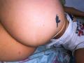 AbbyJanes' big ass with a small black cactus tattoo on the right buttcheek, wearing a white shirt, lying on a bed with a colorful patterned blanket.