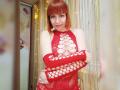 LaylaHottyX with red bob, wearing red lace-up dress and matching red arm sleeves, arms crossed, showing her tits, smiling, standing in front of string lights.
