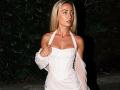 ViktoriiaLu with blonde hair, wearing a sheer white dress, exposing her tits, standing at night, holding dress strap, looking to the side, dark green foliage background.