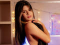 SheylaSpooner with long black hair, glossy pink lips, holding a black cloth over her bare back, looking at the camera with a slight smile, surrounded by warm fairy lights.