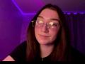 SunnyKlusman with glasses, nose ring, and black shirt, has a slight smile, brown hair, and a purple-lit background. Boobs are not visible. Face is centered in the image.