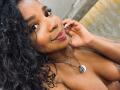 LisaMendez with curly black hair, brown skin, smiling at the camera, wearing a silver necklace with a white circular pendant, touching her cheek, topless, showing her tits.