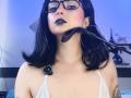 Close-up of CandyParis with black hair, glasses, and black lipstick, wearing a white lace bra, holding a black strap around her neck. Blue-toned background with blurred objects. Slightly sexual tension.