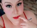 GiiannaMorrys with red lipstick, blue eyes, and pink blush, biting her red-painted finger, wearing red lingerie, showing a tattoo on her right shoulder, looking at the camera.