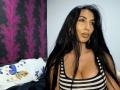 BrunetteBabe69 with long black hair, brown eyes, and big tits in a striped tank top, looking at the camera with a pouty expression. Purple and black wallpaper in background.