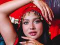 LilyConorr with shiny pink lips, green eyes, and black hair, wearing a gold coin headpiece and red veil, has gold jewelry and manicured nails, posing seductively.