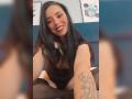 MartinaRey with long black hair, smiling, wearing a black lace top and stockings, showing a tattoo on her arm, sitting on a bed. Boobs are partially visible.