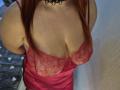 Dinastiia, con lingerie in pizzo rosso e raso, mostra il suo seno grande e formoso in un abito rosso in pizzo con scollatura bassa, indossando una collana nera in pizzo, con i capelli rossi..