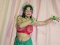 Yurimy with medium tits, red see-through bra, green flower crown, green skirt, and green wristbands, smiling with eyes closed, arms raised, standing against pink curtain.