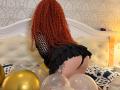 Niferttiti with curly red hair, black fishnet top, and ruffled black skirt, on knees, showing her ass, surrounded by balloons on a white, tufted bed.