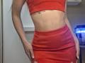 Photograph of a woman, MilaYanis, wearing a tight red crop top and matching red skirt, standing with hands on her hips, highlighting her slim, toned midriff and visible tattoo. Bright lighting.
