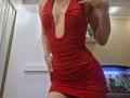 MilaYanis in a tight, red, ruched dress with a deep V-neck, showing her small tits and flat ass. Her hand is on her hip, and she's wearing a necklace.