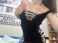 Ramilla in a black, ripped, sleeveless dress with cutouts exposing her tits and sides, hand on chest, brown hair, in a bedroom, with a blue pillow and wooden shelf.