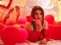 SaraWestt with long brown hair, wearing a red sparkling off-shoulder top and a silver tiara, lies in red balloons, holding a red heart-shaped lollipop, looking at the camera.