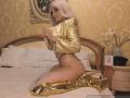MademoiselleBella with platinum blonde wig, gold satin top, black thong, and gold high-heel boots, kneeling on bed, showing her ass and looking back.