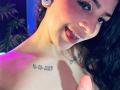 LonxEva with curly black hair, white earbuds, and a tattooed shoulder, sticking out her tongue, and showing a finger to the camera. She has a cute, playful expression.