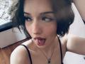 ShanekaKonig has short, wavy brown hair, a nose ring, and a tongue piercing with a ring, wearing a black spaghetti strap top, looking up with her tongue out.
