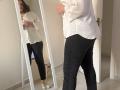 Elliance stands in front of a mirror, wearing a white blouse and black pants, her ass slightly visible, with white sneakers, reflecting her upper body in the mirror.
