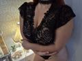 Una donna con seni grandi, coperti di pizzo, braccia incrociate, che indossa una maglia nera in pizzo e un collare, capelli rossi, in piedi, biancheria intima nera, pelle chiara, una lampada e una piccola pianta sullo sfondo..