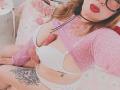 PauleetteRoux with pink sweater, white bra, and white panties, exposing her erect, uncircumcised penis, and a tattoo on her right thigh. She's wearing red lipstick and glasses.