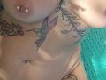 FrenchieLilieFleur's topless torso with a colorful hummingbird tattoo, pierced nipples, and a navel piercing. Her wet skin and floral tattoos are visible.