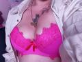 AlessadraJoness' pink lace bra with a red bow, showing her big tits, under a white jacket, with tattoos on her chest and a necklace.