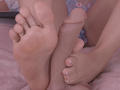 ArianaRogers' feet with a gold ring on the big toe, wrapped around a circumcised, erect penis, showing her painted toenails, on a pink bed.