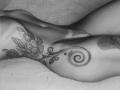 Schwarz-weißes Foto einer nackten Frau, VeronnaInk, die auf dem Rücken liegt und dabei aufwendige Tattoos an ihrer Seite und Hüfte zeigt. Blumen- und spiralförmige Muster. Weiches, sinnliches Licht. Glatte Haut..