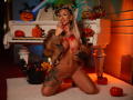 CelineDian, kneeling, nude, with large boobs and tattoos, wearing a fur jacket, touching her lips with red painted nails, surrounded by pumpkins and candles, on a white rug.