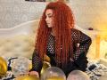 Niferttiti with curly red hair, black fishnet top, and black dress, sitting on bed with gold and silver balloons, hands on balloons, looking down, in a cozy room.