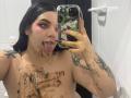 AryButty with chocolate smeared on her tits and tongue out, taking a selfie in a bathroom. She has tattoos, black hair, and pink eyeshadow. Her phone has a green pattern.