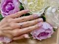 Olivias Hand mit rosafarben lackierten Nägeln, mit einem silbernen Ring mit einem Diamanten, ruht auf weißen und lila Peonienblüten..