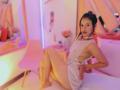 MelisaDaltonn, avec un corps mince et aux courbes prononcées, portant une robe blanche transparente, laissant entrevoir sa poitrine et ses fesses, assise sur un lit, les jambes écartées, dans une pièce éclairée en rose..