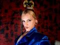 FiorellaNox with blonde hair in a bun, blue velvet jacket, red lipstick, and large ear gauges, looking seductively at the camera. She has a slim face and is sexy.