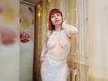 LaylaHottyX with red bob, wearing white lace dress, exposing her tits and pussy, standing by a window with fairy lights and floral decor, looking confident.