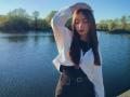 EleanoraMirabal stands by a lake, wearing a white shirt and black high-waisted pants, with one hand in her brown hair, eyes closed, and a calm expression.