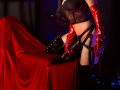 AfroditaDoir with red latex gloves, black lace bra, and strappy black harness, kneeling on a red velvet-covered table, holding a black leather belt. Boobs visible.