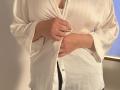Elliance's hands are buttoning a white, silky shirt that's slightly open, revealing a hint of her tits. She's in a bright room with a window.