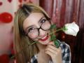 Kaiix with blonde hair, black glasses, and red lipstick, holding a white rose between her teeth, smiling, wearing a black-and-white checkered shirt.