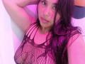 GemmaBellini with pinkish-purple lighting, long black hair, wearing a black fishnet top that reveals her large tits, arm raised, looking at the camera with a slight smile.