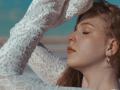 StellaLane with fair skin and brown hair, eyes closed, wearing a white lace long-sleeve top, arms raised, small silver hoop earrings, soft sunlight on her face, serene expression.