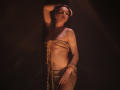 TriciaBarnes with her tits covered in rough rope, wearing a tiny metallic skirt, standing in dark shadow, arm above head, looking seductive, her belly button pierced.