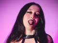 KendraSimons has black lipstick, long dark hair, and is sticking out her tongue with eyes half-closed, wearing a black choker, against a pink gradient background.