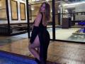 AvaQueen with blonde hair, black dress, and high heel boots, showing her ass and back, standing by a pool, looking over her shoulder, smiling.