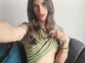 LunarisSmith, with long brown hair, green top, and pink lipstick, is lifting their shirt to show a small, perky breast. Wearing bracelets, sitting on a blue couch.
