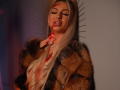 CelineDian with long blonde hair, wearing a furry coat, blood on her face, and hands near her tits, has a sexy, sultry expression with her eyes closed.