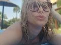 MissFrenchy with wet blonde hair, wearing dark sunglasses, pouting her pink lips, showing her bare shoulders, and slightly squinting. She's outdoors, with a blurred background.