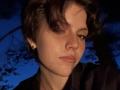 HanhFontillas with short, curly brown hair, freckled face, small nose piercing, and black shirt, looking slightly to the side, against a dark blue background.
