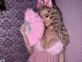 MademoiselleBella with pink fluffy fan, holding it near her tits, wearing pink dress with fluffy trim, pink bow, and jewelry, long curly blonde hair, pink lipstick, and eyeliner.