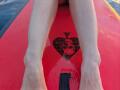 AuroraStar's legs are spread on a red surfboard with a black heart and diamond pattern, her bare feet gripping the wet surface.